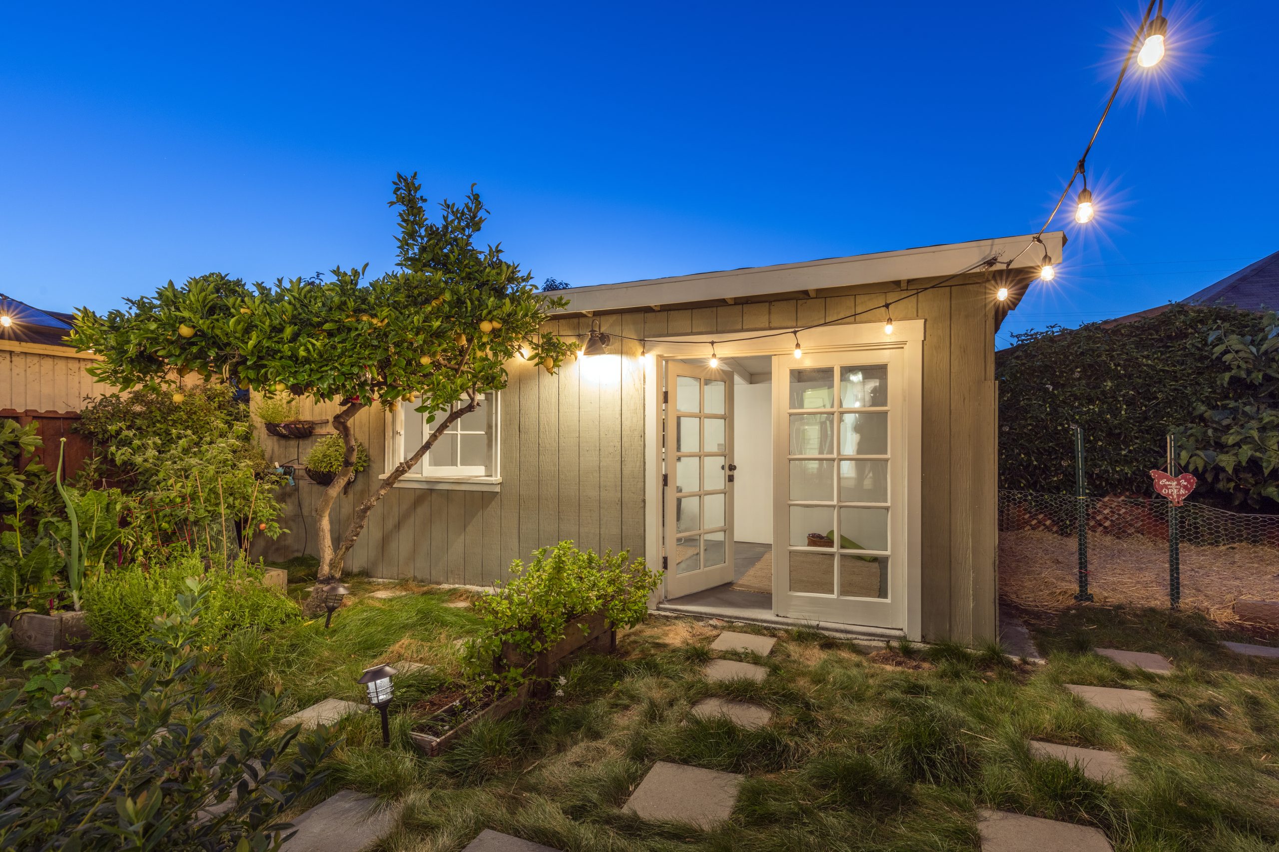 An Acessory Dwelling Unit, or ADU for short, with beige wooden boards for walls, a white framed window and a white framed glass door, with a yard in front containing overgrown grass, square stepping tones, plants and a lemon tree with a few fruits hanging.